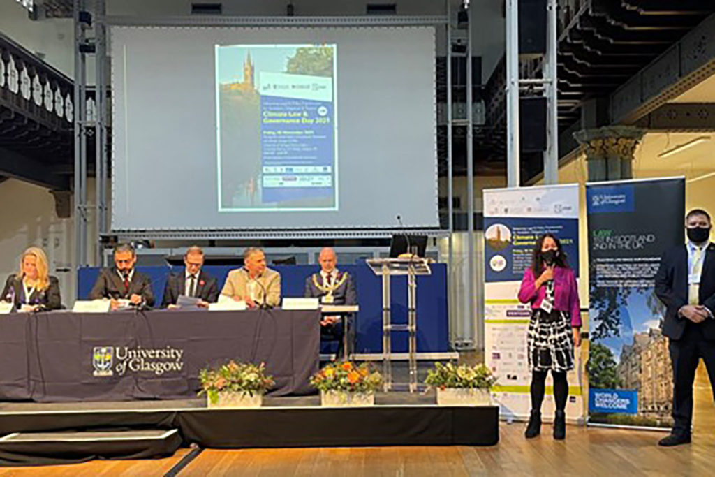 Four males and one female sit a table with University of Glasglow tablecloth. Female speaker stands beside the panel table holding a microphone