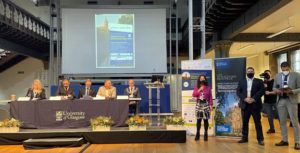 Four males and one female sit a table with University of Glasglow tablecloth. Female speaker stands beside the panel table holding a microphone