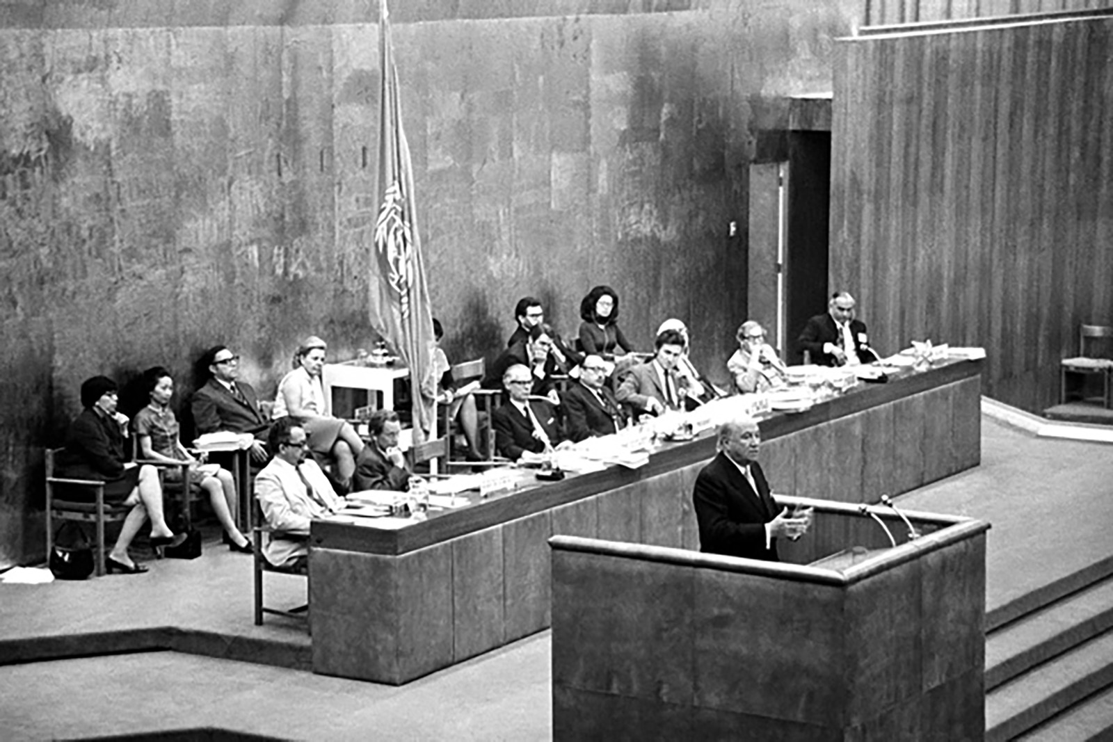 1 male at a podium with 8 people seated behind a long desk and 6 people seated behind in chairs. The UNCTAD flag is on a flag pole between the people seated.