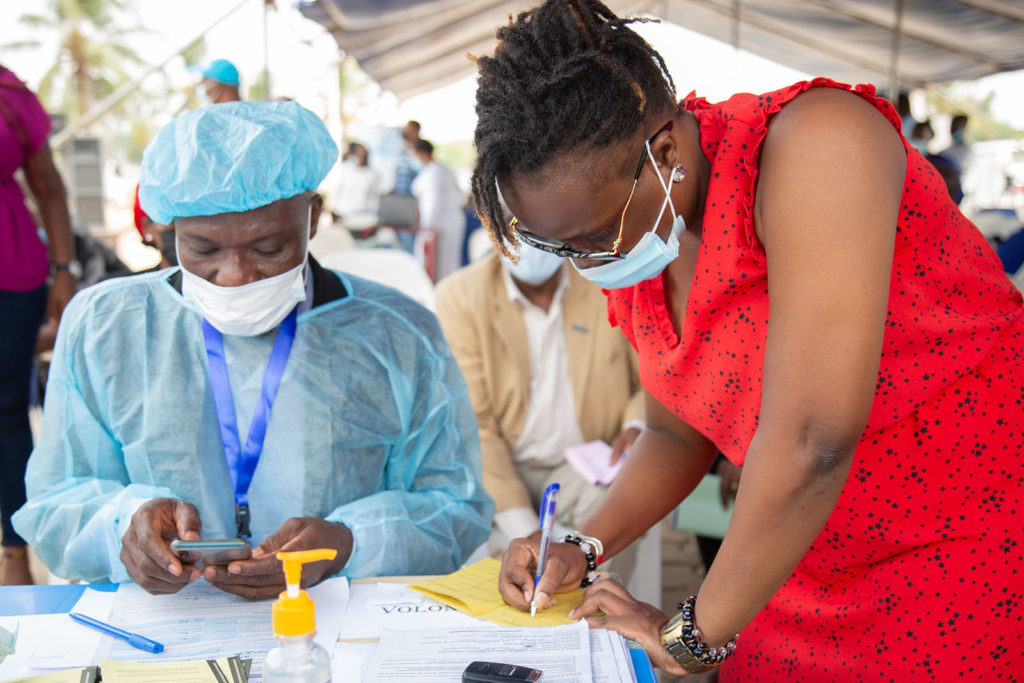 Male seated with a medical cap, gown and mask, at a table. A woman is wearing a medical mask making a note on a piece of paper