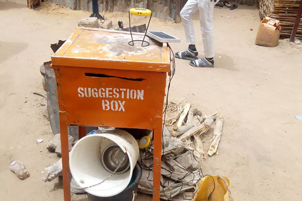 A metal box on legs with a slot in the front and the words "Suggestion Box". Debris and a bucket are under the box.