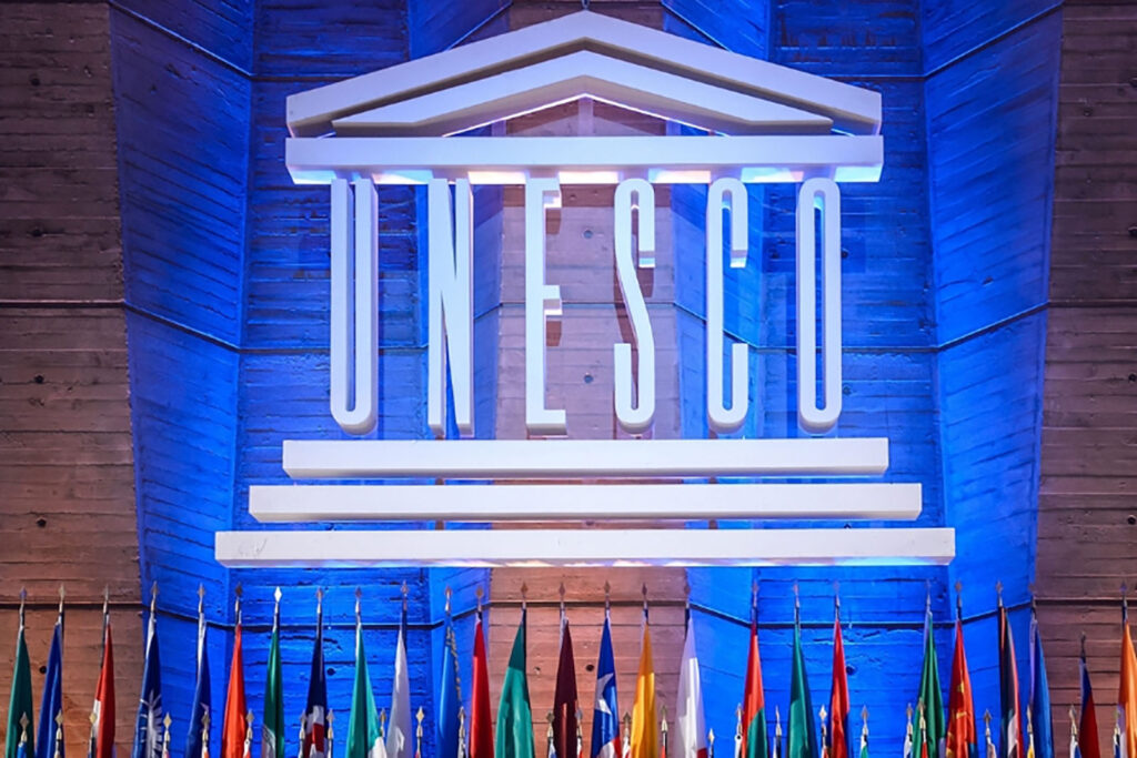 UNESCO logo that has the term UNESCO with three lines below and a roof above the name. The logo is hung in front of a blue background with a row of flags from different countries below.