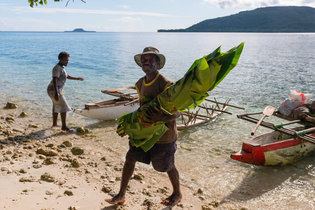 A man carrying banana leaves near the edge of water with a women and a boat in the background