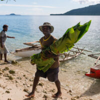 A man carrying banana leaves near the edge of water with a women and a boat in the background