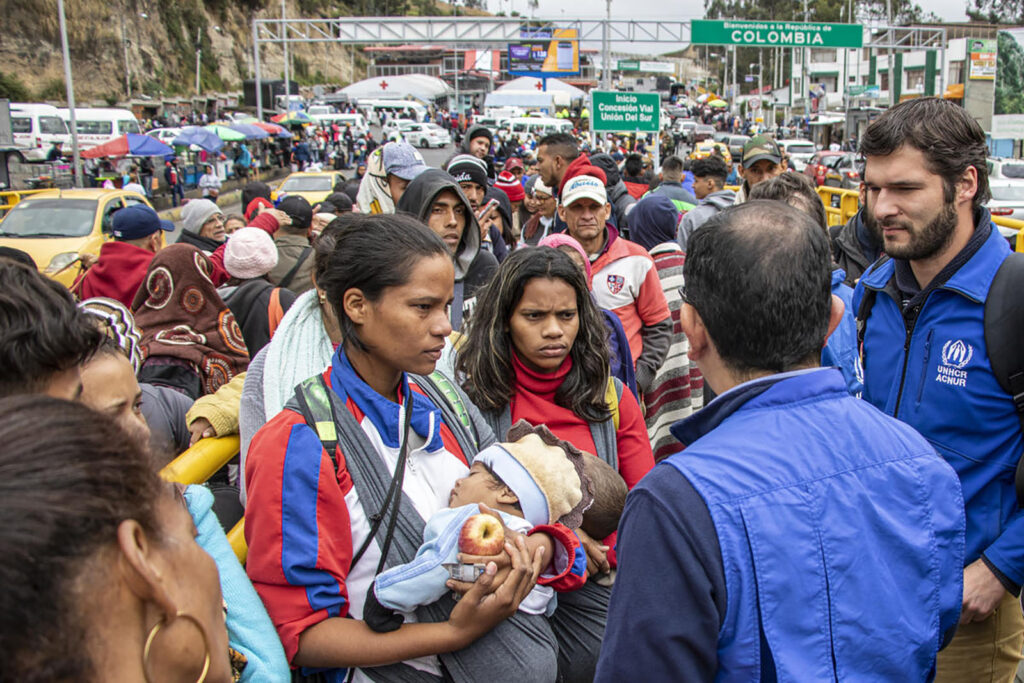 A woman in a crowd, holding a baby and speaking to a man in a blue vest