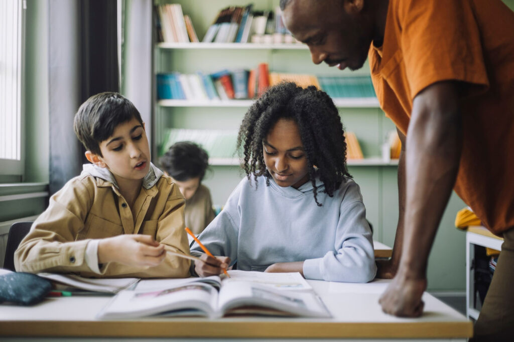 Two children working at a desk with a male teacher looking at their work.