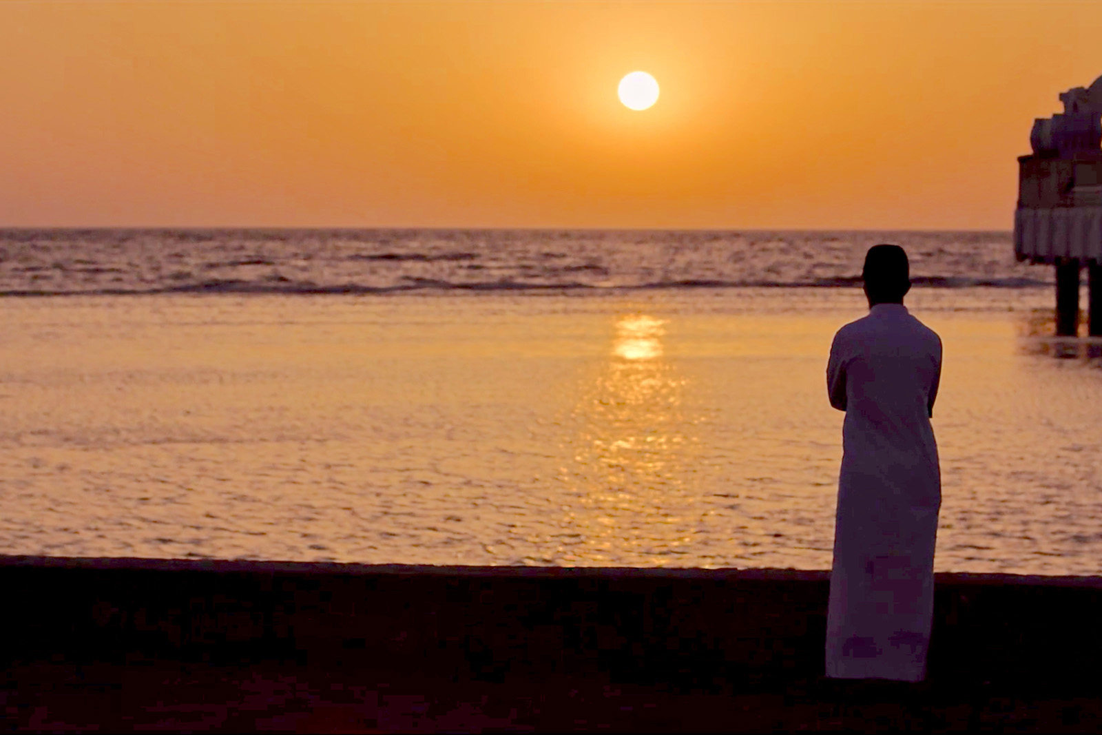 Silhouette of a man standing on a beach looking at the water and the sun setting in the sky.