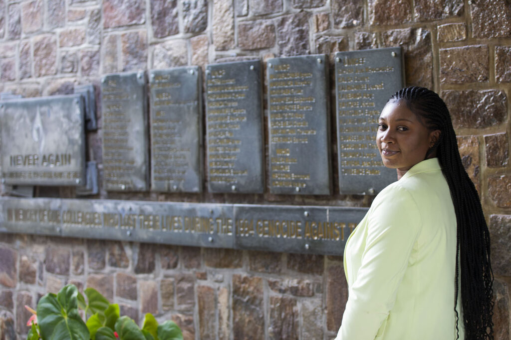 Adwoa standing next to a wall with plaques displaying names and the words, "never again"