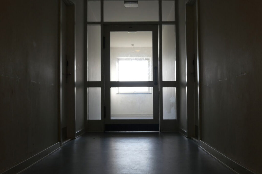 A dark corridor with a glass door. A window with light shining in is behind the glass door.