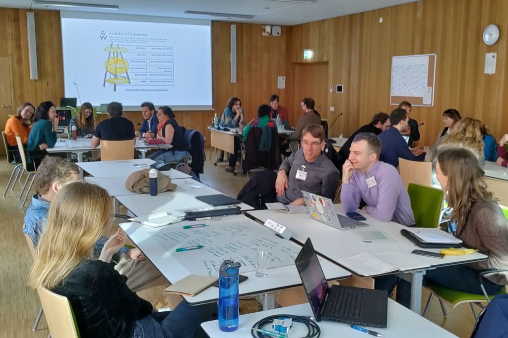 Groups of people sitting at tables with a presentation on a screen at the front of the room.