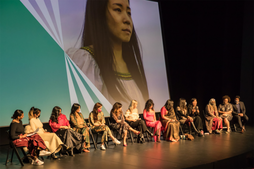 A row of people sitting in chairs on a stage with a screen behind them. A young woman is on the screen looking forward with wind blowing her hair from behind.