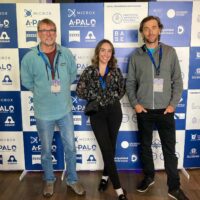 Hope Tracey standing with two colleagues in front of a banner with logos