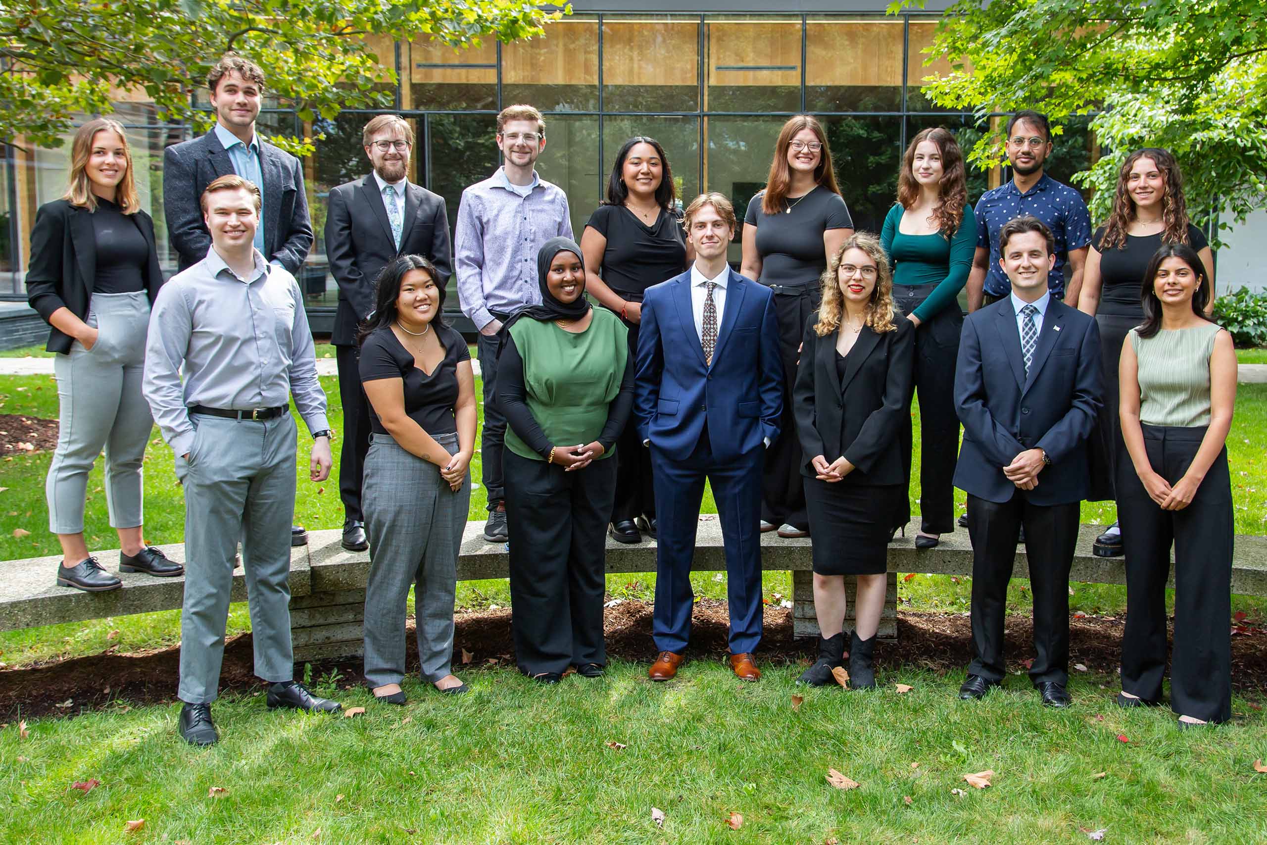 16 people standing in two rows on grass and a bench in front of a building and a tree