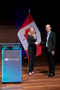 Ann and Benjamin standing on a stage shaking hands. The Canada flag is behind them and a podium with the ONHcloud logo is beside them.